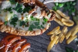 A close-up of a dish featuring a crispy base topped with melted cheese and fresh parsley. Slices of ripe tomato with a drizzle of white sauce sit beside golden-brown fries. The background includes scattered herbs on a textured surface.