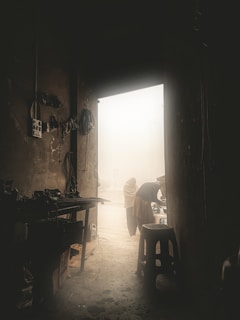 An interior view of a dark workshop or garage with various tools and equipment along the left wall. An open door reveals a foggy or dusty outdoor environment where two people are seen, one with a hood bending over and another standing near them. The light outside contrasts with the dimly lit interior, creating a dramatic contrast.