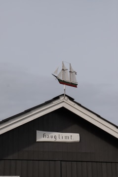 A black wooden building with a triangular roof features a small model of a three-masted sailing ship on its peak. Below the ship, there is a sign with the word 'havglimt'. The sky is overcast, providing a muted backdrop.