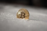 A gold-colored Bitcoin coin is partially embedded in snow, glistening under light, providing a stark contrast between the metallic sheen and the soft texture of the snow.