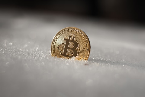 A gold-colored Bitcoin coin is partially embedded in snow, glistening under light, providing a stark contrast between the metallic sheen and the soft texture of the snow.