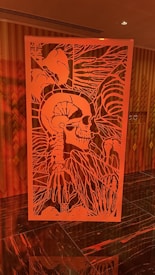A detailed and intricate red metal art piece resembling a human skeleton is prominently displayed. The artwork includes outlines of bones and abstract patterns surrounding the skull and ribcage. The background features a wooden panel wall with vertical patterns, creating a contrast with the art piece.