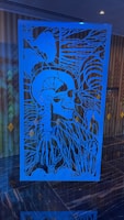 An intricate blue art piece with a skull and rib cage, surrounded by abstract plant-like lines and patterns. It is placed in front of a patterned wall with muted tones.