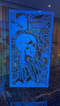 An intricate blue art piece with a skull and rib cage, surrounded by abstract plant-like lines and patterns. It is placed in front of a patterned wall with muted tones.