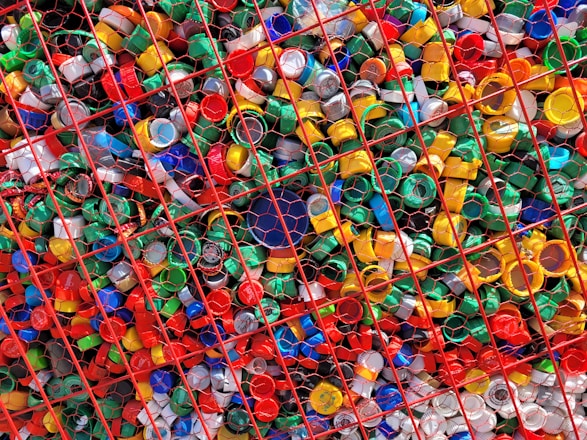 A large collection of colorful plastic bottle caps is contained within a red metal grid. The caps vary in color, including shades of red, green, yellow, blue, and white. They are densely packed together, creating a vibrant and chaotic visual effect.