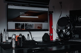 A desktop setup featuring a large monitor displaying the Unsplash homepage, surrounded by various electronic accessories. The desk is cluttered with headphones placed on a stand, a keyboard, mouse, and several bottles. A gaming PC with visible hardware details is situated to the side, and the lighting is dim, creating a moody atmosphere.