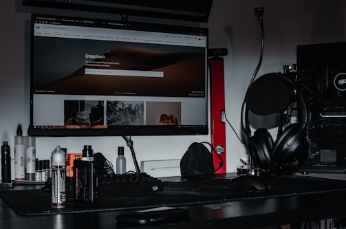 A desktop setup featuring a large monitor displaying the Unsplash homepage, surrounded by various electronic accessories. The desk is cluttered with headphones placed on a stand, a keyboard, mouse, and several bottles. A gaming PC with visible hardware details is situated to the side, and the lighting is dim, creating a moody atmosphere.