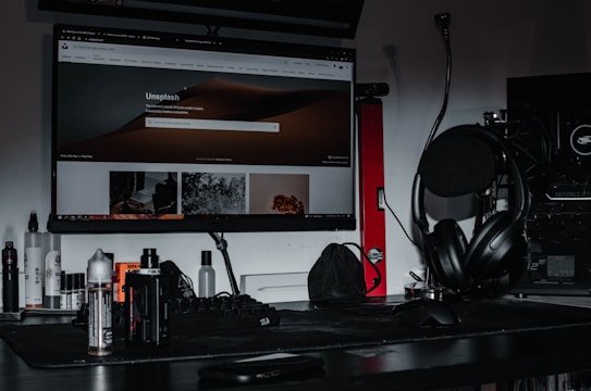 A desktop setup featuring a large monitor displaying the Unsplash homepage, surrounded by various electronic accessories. The desk is cluttered with headphones placed on a stand, a keyboard, mouse, and several bottles. A gaming PC with visible hardware details is situated to the side, and the lighting is dim, creating a moody atmosphere.