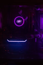 A dimly lit computer interior showcasing a purple LED-lit CPU cooler labeled 'Deep Cool' and a visible GeForce GTX graphics card with blue lighting. The components are encased within a dark chassis and connected with cables.