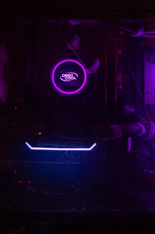 A dimly lit computer interior showcasing a purple LED-lit CPU cooler labeled 'Deep Cool' and a visible GeForce GTX graphics card with blue lighting. The components are encased within a dark chassis and connected with cables.