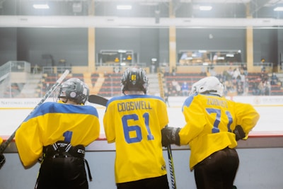 Three young hockey players wearing yellow and blue jerseys with helmets are standing at the edge of a hockey rink. They are preparing to enter the ice, holding hockey sticks. The rink is indoors, with empty spectator seats visible in the background.