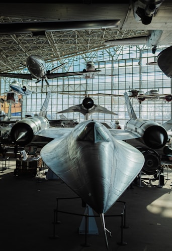 A large, sleek, black aircraft is prominently displayed in an indoor museum setting, surrounded by other aircraft. The environment is well-lit with natural light streaming through large glass windows, creating a bright and airy atmosphere.