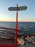 A signpost with two directional arrows pointing towards 'Inciraltı 1km' and 'Konak 9km' is situated on a seaside path. The sign is mounted on a red pole, matching the red railing to the left. The background features a calm sea under a clear sky, with the sun setting, casting a warm glow over the rocks along the shoreline.