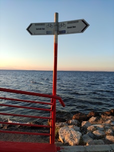 A signpost with two directional arrows pointing towards 'Inciraltı 1km' and 'Konak 9km' is situated on a seaside path. The sign is mounted on a red pole, matching the red railing to the left. The background features a calm sea under a clear sky, with the sun setting, casting a warm glow over the rocks along the shoreline.