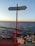 A signpost with two directional arrows pointing towards 'Inciraltı 1km' and 'Konak 9km' is situated on a seaside path. The sign is mounted on a red pole, matching the red railing to the left. The background features a calm sea under a clear sky, with the sun setting, casting a warm glow over the rocks along the shoreline.