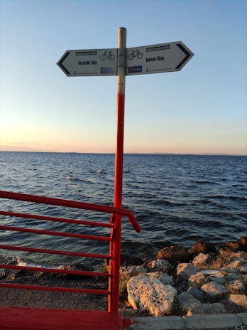 A signpost with two directional arrows pointing towards 'Inciraltı 1km' and 'Konak 9km' is situated on a seaside path. The sign is mounted on a red pole, matching the red railing to the left. The background features a calm sea under a clear sky, with the sun setting, casting a warm glow over the rocks along the shoreline.