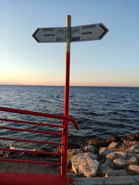 A signpost with two directional arrows pointing towards 'Inciraltı 1km' and 'Konak 9km' is situated on a seaside path. The sign is mounted on a red pole, matching the red railing to the left. The background features a calm sea under a clear sky, with the sun setting, casting a warm glow over the rocks along the shoreline.