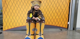 A child is dressed in traditional attire which includes a fur hat, an ornate coat with intricate patterns, and oversized yellow boots adorned with blue details. The background features a yellow, diamond-patterned design.