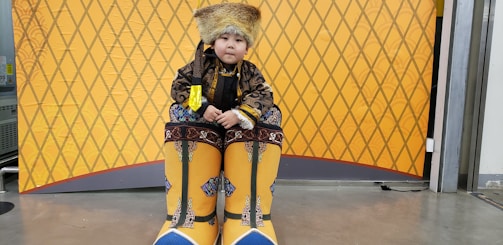 A child is dressed in traditional attire which includes a fur hat, an ornate coat with intricate patterns, and oversized yellow boots adorned with blue details. The background features a yellow, diamond-patterned design.