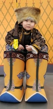 A young child dressed in traditional attire, including a large fur hat and ornate oversized boots with intricate patterns. The background features a geometric design in warm colors.