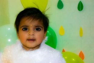A young child with dark hair, wearing a fluffy white outfit, stands in front of a backdrop with colorful balloons and paper cut-out shapes resembling leaves in yellow, green, and orange. The background is light green, complementing the decorations.