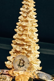 A golden Christmas tree ornament stands beside several gold-colored Bitcoin replicas. The scene is set against a dark background, highlighting the shiny and reflective surfaces of the coins and ornament.