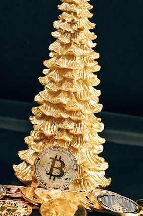 A golden Christmas tree ornament stands beside several gold-colored Bitcoin replicas. The scene is set against a dark background, highlighting the shiny and reflective surfaces of the coins and ornament.