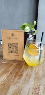 A wooden table holds a glass of a yellow citrus drink filled with ice and garnished with orange slices and a sprig of rosemary. Beside the drink is a small decorative plant in a dark vase. A stand-up card display provides information and a QR code, indicating the name 'coffeenatics'. The background is a plain gray wall.