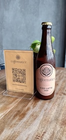 A brown glass bottle labeled 'Coffeenatics Iced Latte Okinawa' is sitting on a wooden table. Next to it is a stand with a card featuring a QR code and the brand 'Coffeenatics' logo. In the background, there is a small potted plant with green leaves against a gray wall.
