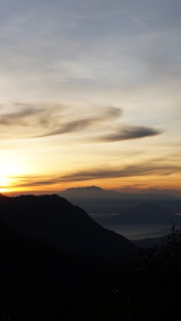 A serene mountain landscape at sunrise with silhouetted hills in the foreground and layers of mountains receding into the distance. The sky is painted with warm hues of orange and yellow, transitioning into soft blues and grays as it extends upward.