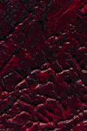 Close-up of a textured dark canvas with streaks of deep crimson and black.