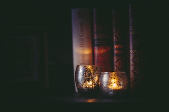 Dimly lit, ornate candle holders with flickering flames are placed in front of a row of thick, decorative hardcover books. The scene creates a cozy and intimate atmosphere with soft shadows.