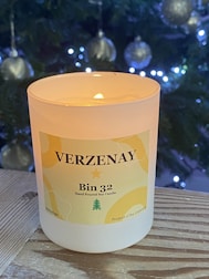 A lit Verzenay Bin 32 hand-poured soy candle is situated on a wooden surface. In the background, a Christmas tree adorned with silver baubles and glowing lights adds a festive atmosphere.
