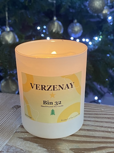 A lit Verzenay Bin 32 hand-poured soy candle is situated on a wooden surface. In the background, a Christmas tree adorned with silver baubles and glowing lights adds a festive atmosphere.