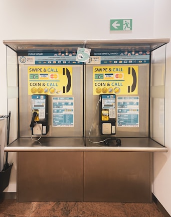 A pair of public payphones are mounted on a wall inside an enclosed booth with glass panels. The payphones offer options for swiping a card or inserting coins to make calls. Above, there are instructions and advertisements for international phone calls. A green emergency exit sign is visible on the wall.