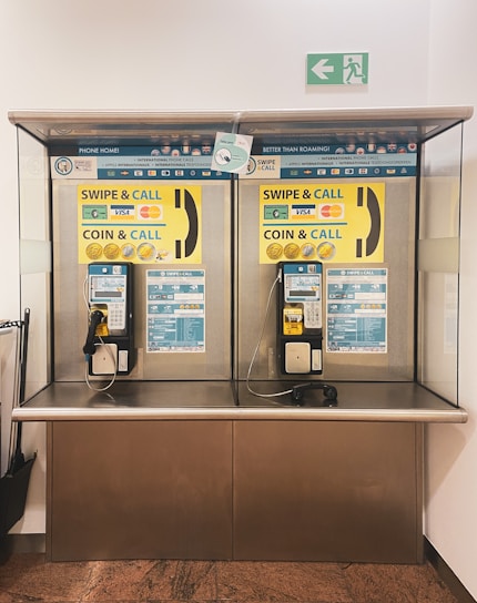 A pair of public payphones are mounted on a wall inside an enclosed booth with glass panels. The payphones offer options for swiping a card or inserting coins to make calls. Above, there are instructions and advertisements for international phone calls. A green emergency exit sign is visible on the wall.