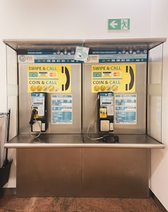 A pair of public payphones are mounted on a wall inside an enclosed booth with glass panels. The payphones offer options for swiping a card or inserting coins to make calls. Above, there are instructions and advertisements for international phone calls. A green emergency exit sign is visible on the wall.