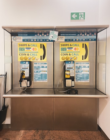 A pair of public payphones are mounted on a wall inside an enclosed booth with glass panels. The payphones offer options for swiping a card or inserting coins to make calls. Above, there are instructions and advertisements for international phone calls. A green emergency exit sign is visible on the wall.