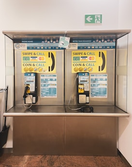 A pair of public payphones are mounted on a wall inside an enclosed booth with glass panels. The payphones offer options for swiping a card or inserting coins to make calls. Above, there are instructions and advertisements for international phone calls. A green emergency exit sign is visible on the wall.