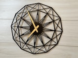 A wall clock with a geometric design featuring a circular layout and intricate black lines forming a pattern. The clock hands are gold and contrast with the black framework. It is mounted on a light wooden or laminated surface, contributing to a modern and minimalistic aesthetic.