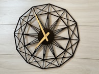 A wall clock with a geometric design featuring a circular layout and intricate black lines forming a pattern. The clock hands are gold and contrast with the black framework. It is mounted on a light wooden or laminated surface, contributing to a modern and minimalistic aesthetic.