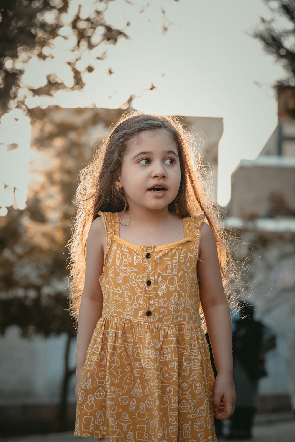 a little girl in a yellow dress standing in front of a tree