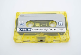 A vintage audio cassette tape with a transparent yellow case and a gray label indicating 'KAMICHI Low Noise High Output' for side B. The cassette tape is labeled with 'LH C90' and features visible reels and spools inside the clear casing.