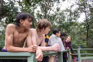 A group of young people are gathered on a wooden railing in an outdoor setting surrounded by trees. They appear relaxed, with some wrapped in towels. The atmosphere is casual and the individuals are engaged in conversation, suggesting a social or leisure activity.