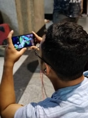 A person is seated, holding a smartphone horizontally while watching a video or playing a game. The individual is wearing glasses and has red earphones plugged in. The background includes a blurred figure and a dimly lit environment.