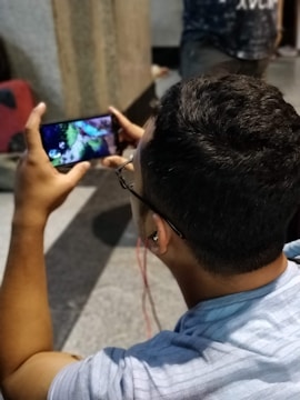A person is seated, holding a smartphone horizontally while watching a video or playing a game. The individual is wearing glasses and has red earphones plugged in. The background includes a blurred figure and a dimly lit environment.