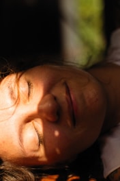 A person with eyes closed is basking in sunlight, creating a peaceful ambiance. The sunlight casts soft shadows across the face, highlighting the tranquil expression and smooth skin.