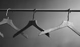 Three wooden clothes hangers are suspended from a horizontal metal rod. The background is a plain, neutral wall, and the image is presented in black and white, emphasizing the textures and shapes of the hangers.