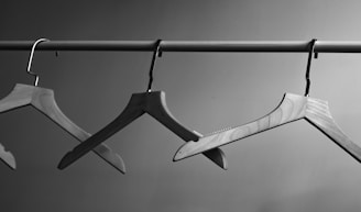 Three wooden clothes hangers are suspended from a horizontal metal rod. The background is a plain, neutral wall, and the image is presented in black and white, emphasizing the textures and shapes of the hangers.
