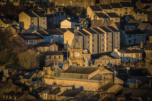 A quaint townscape featuring closely packed stone buildings dominated by a prominent clock tower. The buildings vary in size and style, with some modern apartments in the background. Sunlight casts a warm glow over the scene, enhancing the textures and colors of the architecture.